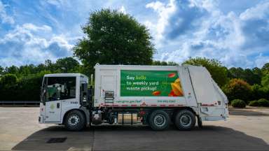 A trash truck with the words "Say hello to weekly yard waste" on it sits parked in a lot with blue skies, clouds and trees as the backdrop.