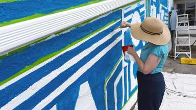 A woman in a straw hat is painting blue lines on the side of a building.