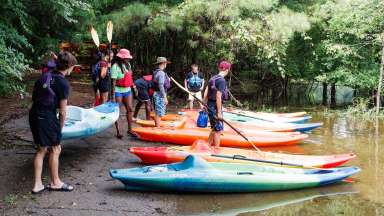 teenagers on shore of a lake holding paddles. Kayaks are lined up on shore