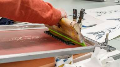 person uses a squeegee to pull ink through a screenprinting screen depicting a dog