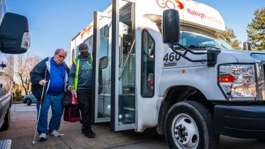 A man in a yellow vest helps a man with a cane get on an access transit vehicle.