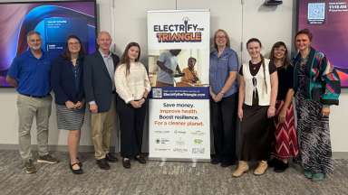 A group of people standing beside an Electrify the Triangle banner featuring program details and partner logos.