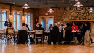 A group of people sit in a circle on folding chairs in a large room, listening to a woman leading a discussion.