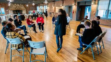 A group of people sit in a circle on folding chairs in a large room, listening to a woman standing in the center leading a discussion.