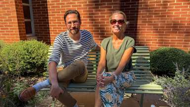 Podcast hosts site on a bench with a brick building behind them and greenery around the bench