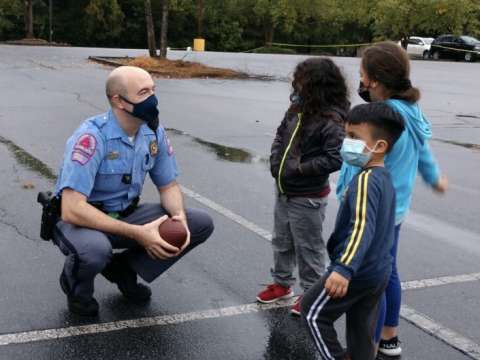 Officer with a football kneels down in parking lot facing 3 children