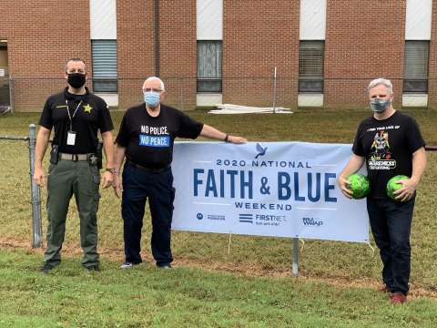 3 men facing camera standing besides a faith and blue banner outside