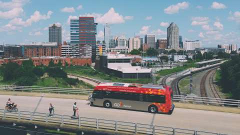 An electric bike, electric scooters and an electric GoRaleigh transit bus in front of the Raleigh City skyline.