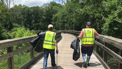 2 people in safety vests with bags and litter grabbers walking on a bridge