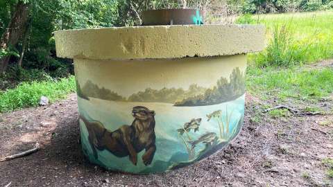 A mural featuring an otter and fish is painted on the side of a sewer riser