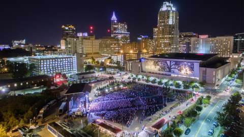 Aerial image of red hat amphitheater at night full of people with Convention Center and Raleigh Skyline in background.