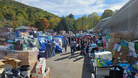 People picking up goods from a huge distribution area full of supplies. Fall colors show in the trees of the mountains in the background.