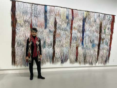 Artist, a slight, white woman with short grey hard, in front of a textured fiber wall hanging in a museum.