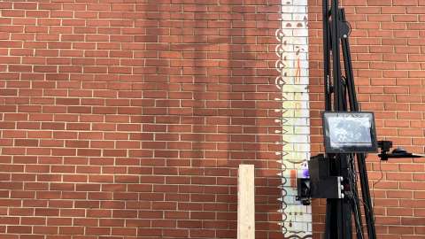 Tall black printer applying white paint to a brick wall to create a mural.