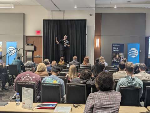 Photo of a presenter at the conference, showing them speaking on the stage and the backs of the attendees