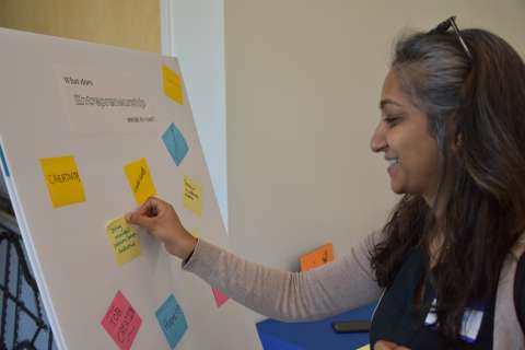 Woman smiling and placing a sticky note on a board that says &quot;What does entrepreneurship mean to you?&quot;