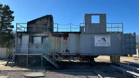 A one-story raised metal structure with stairs leading up to entrance. Burned metal can be seen on parts of the structure.
