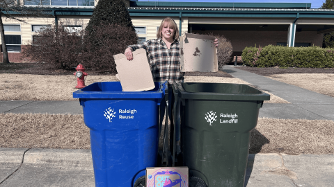 A woman holds a pizza box over a trash bin and a recycling bin to show that the top of a pizza box can be recycled, but the bottom cannot.