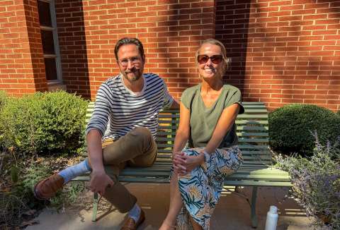 Podcast hosts site on a bench with a brick building behind them and greenery around the bench
