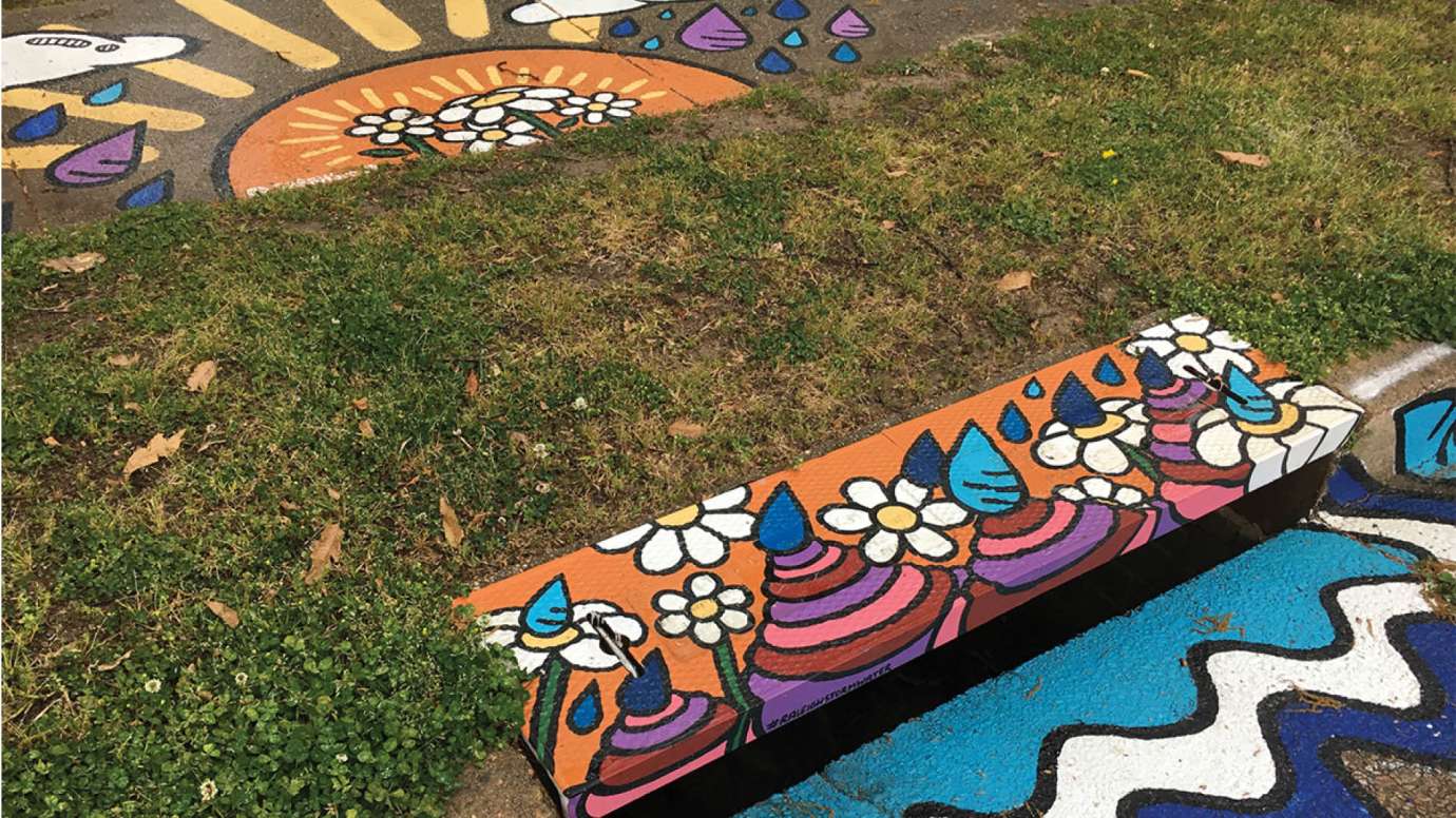 A storm drain and sidewalk mural on Peterson Street with a big yellow sun and pink, purple, and blue raindrops