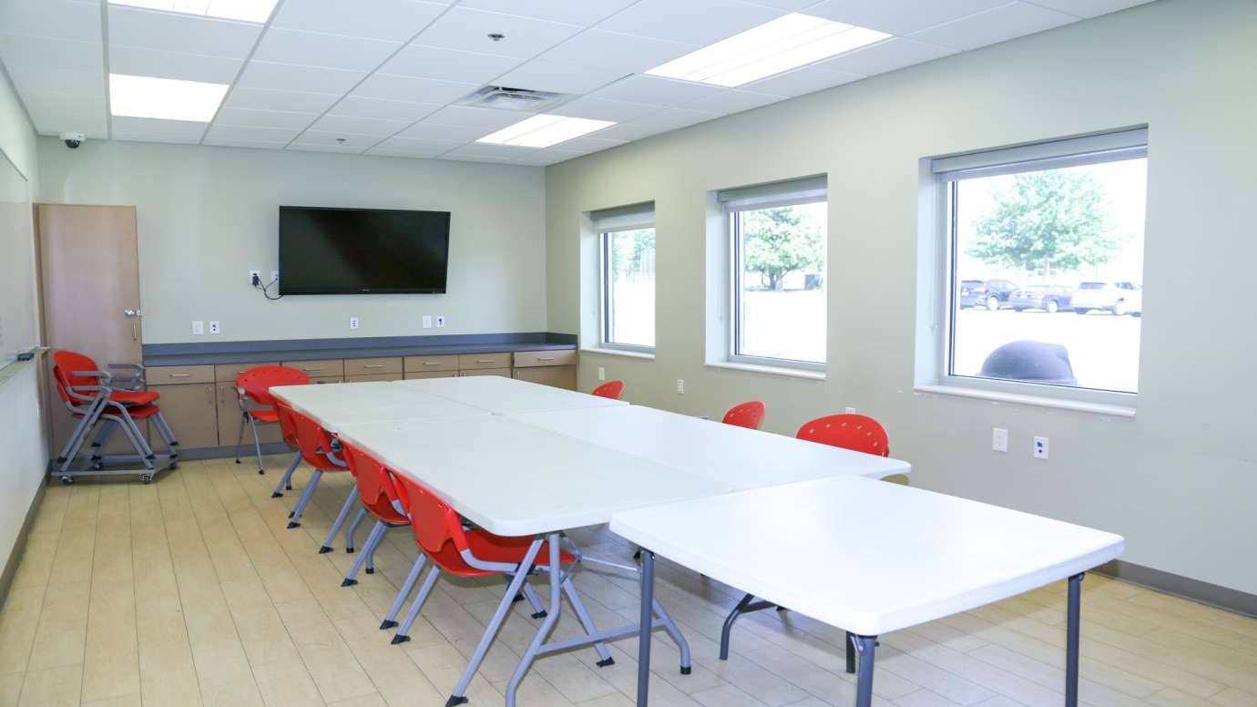 A classroom with a long table, chairs and a mounted TV