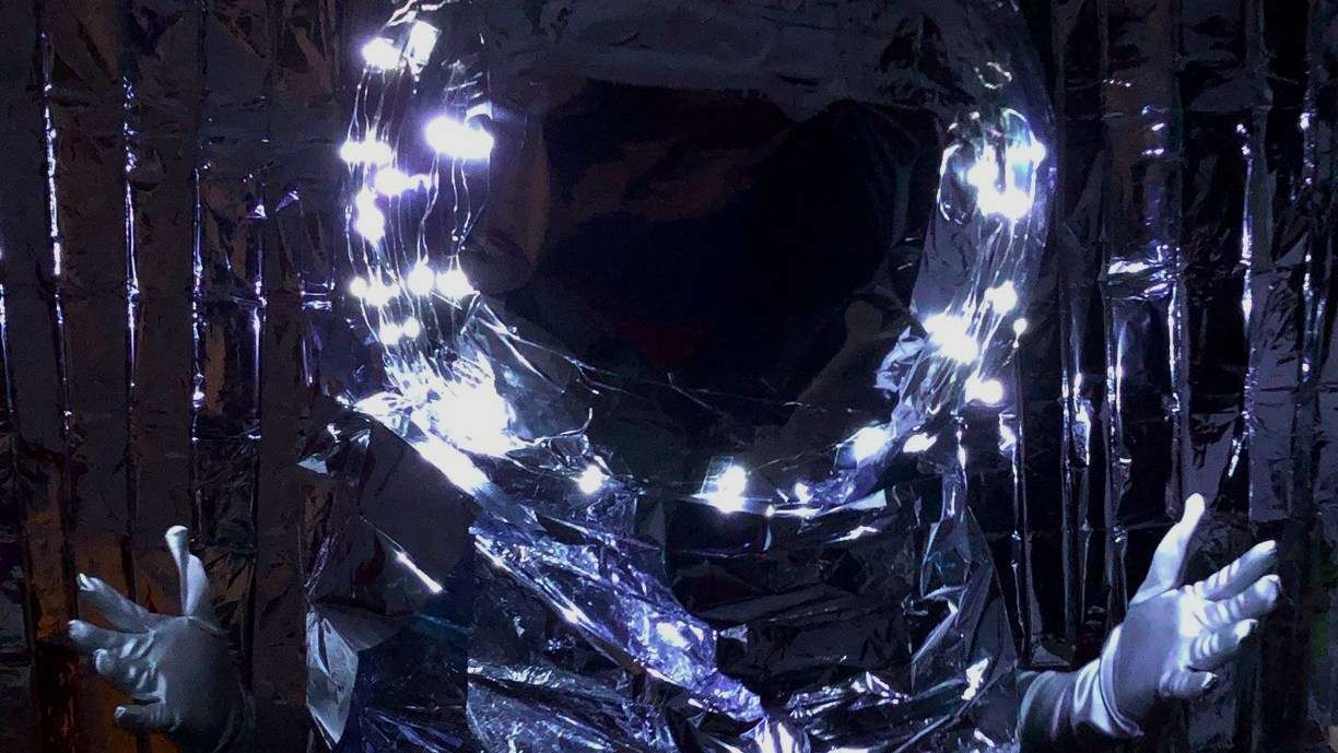 A person covered in silver, shiny material wearing a large spherical silver helmet posing with their arms open towards the camera.