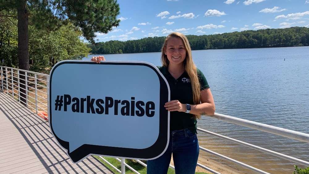 Girl smiling holding Parks Praise sign in front of lake