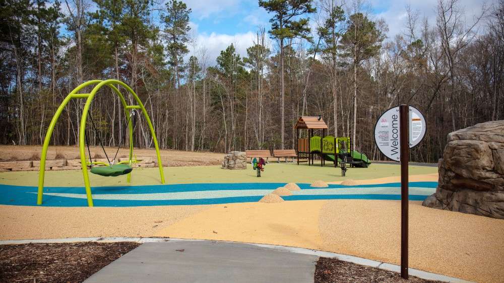 Sidewalk leads to welcome sign at playground with rubberized surface and swings on left and climbing boulder on right