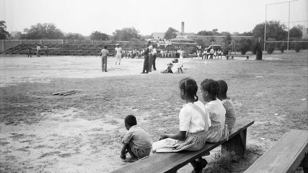 Black and white image of kids sitting on bleachers in forefront with baseball playing in background