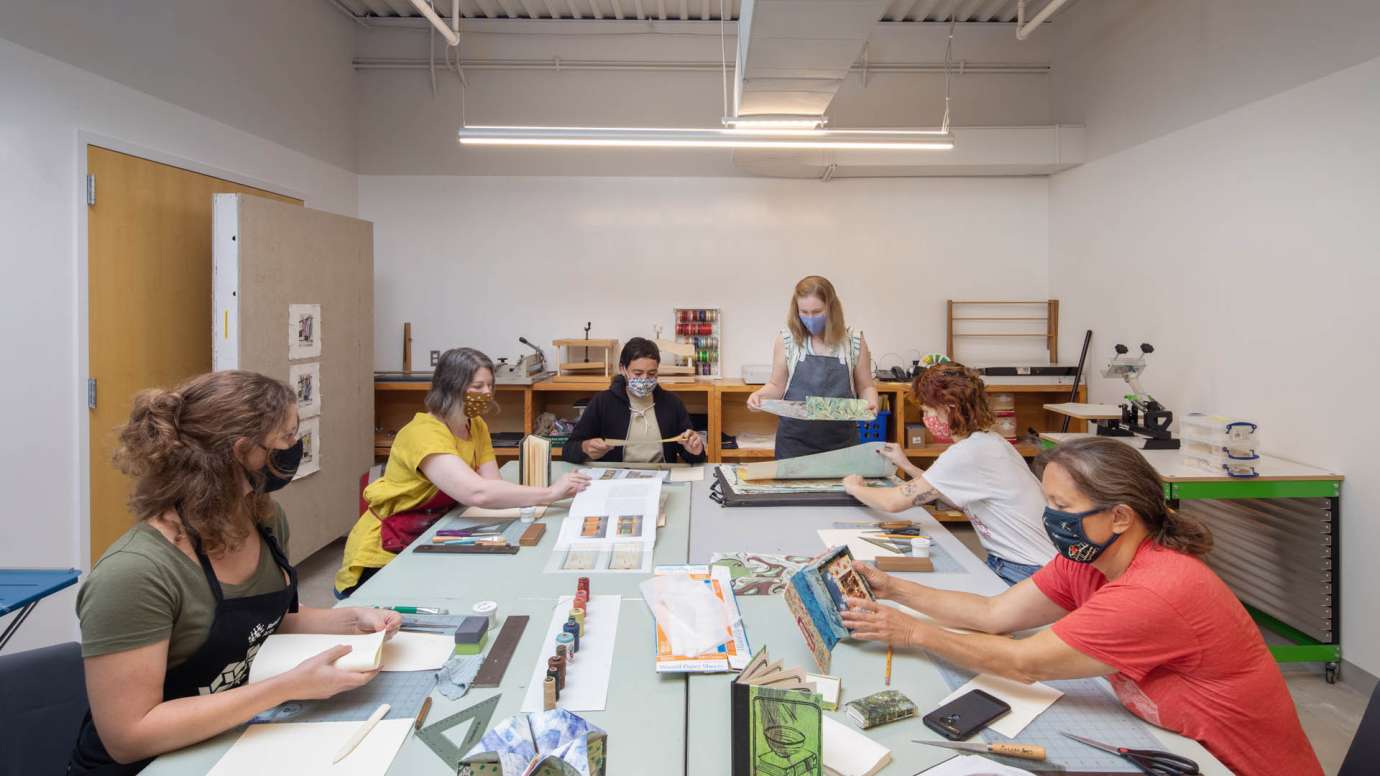 A group of people sit at a table as they work with materials in the Bookmaking & Printmaking studio at Pullen Arts Center