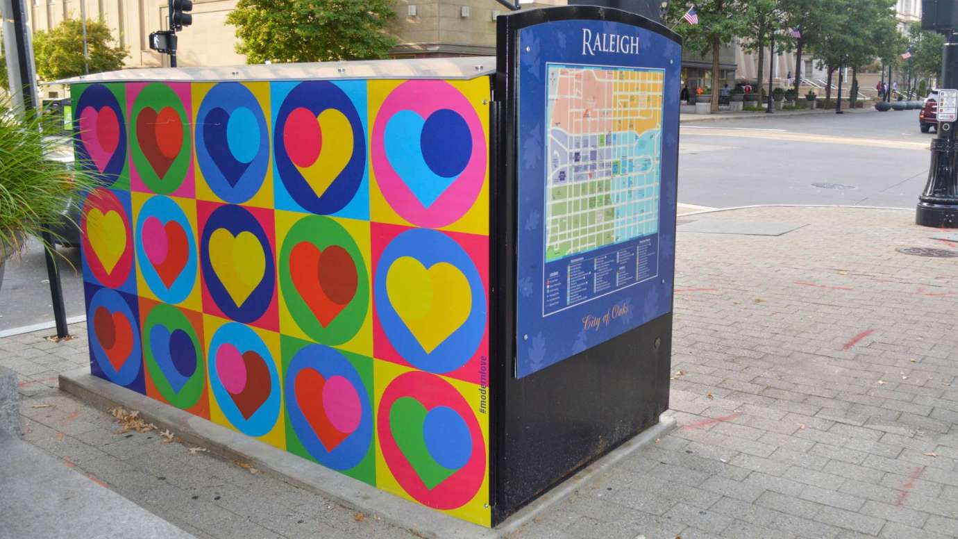 Artwork on a news rack kiosk that features a grid of colorful squares that contain colorful hearts, within some of the hearts is a colorful circle