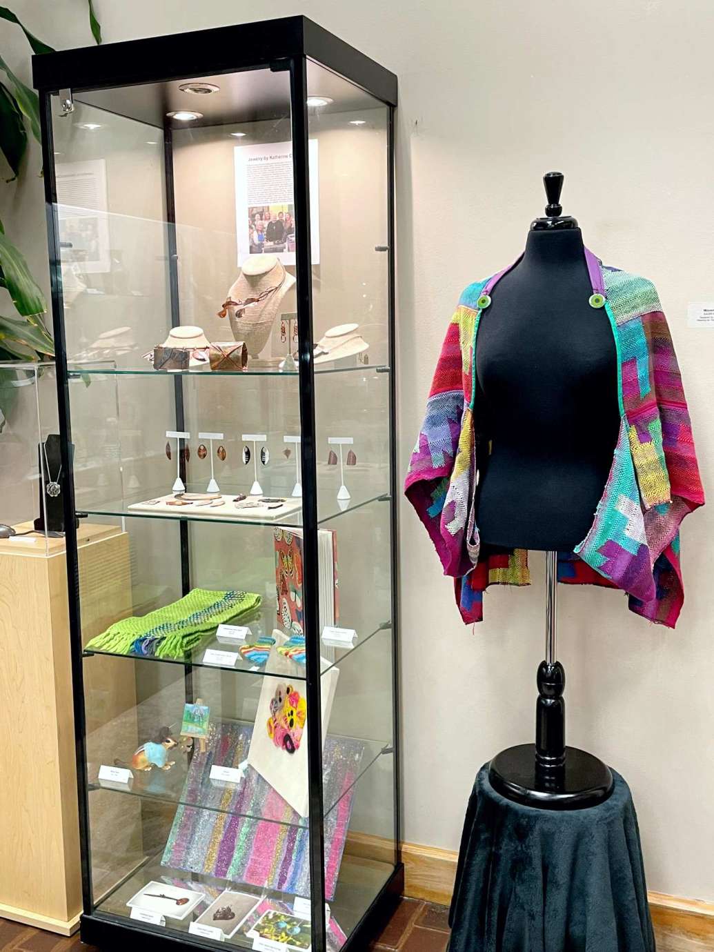 A display case filled with locally made jewelry is seen on left next to a woven textile shawl/jack on a mannequin on the right. 