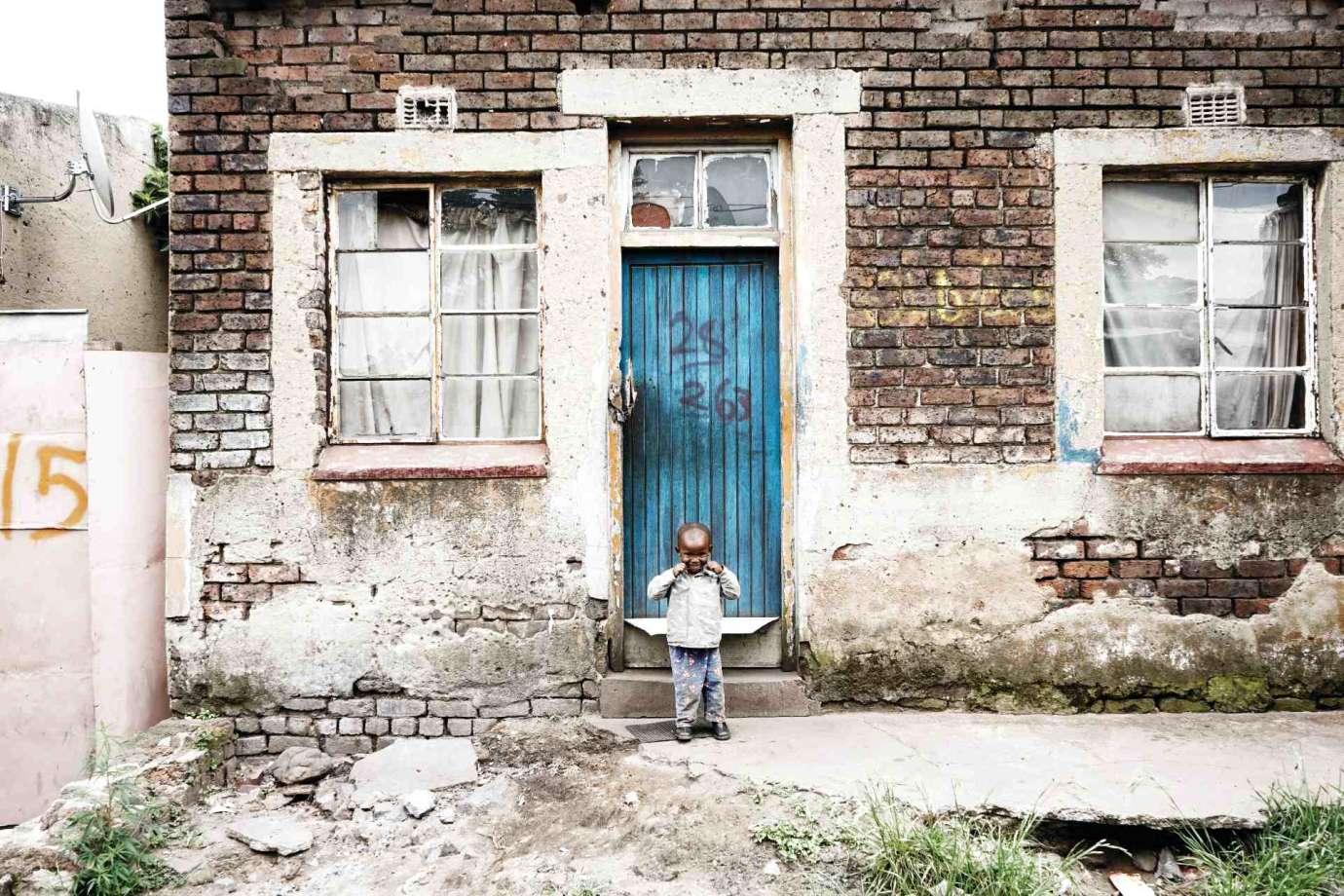 A photograph of a small boy standing in front of a blue door of a brick building by artists Samantha Everette