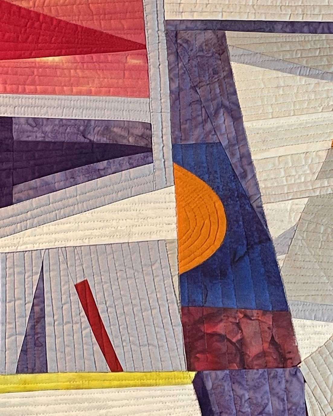 A quilt with blue, red, purple, gray, and yellow abstract lines, triangles, rectangles and a bright orange half circle.