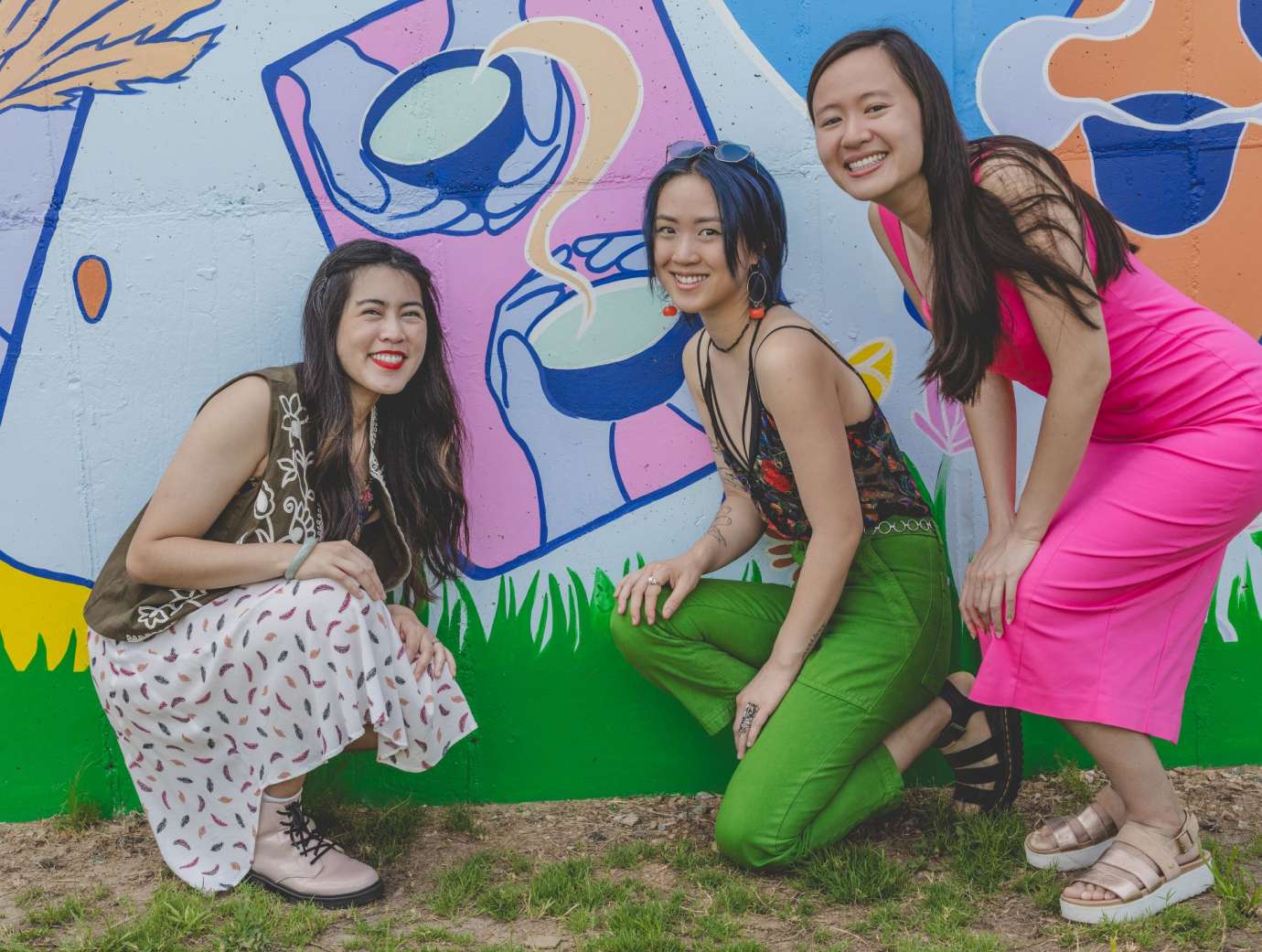 Three young Asian American women kneeling in front of a colorful mural