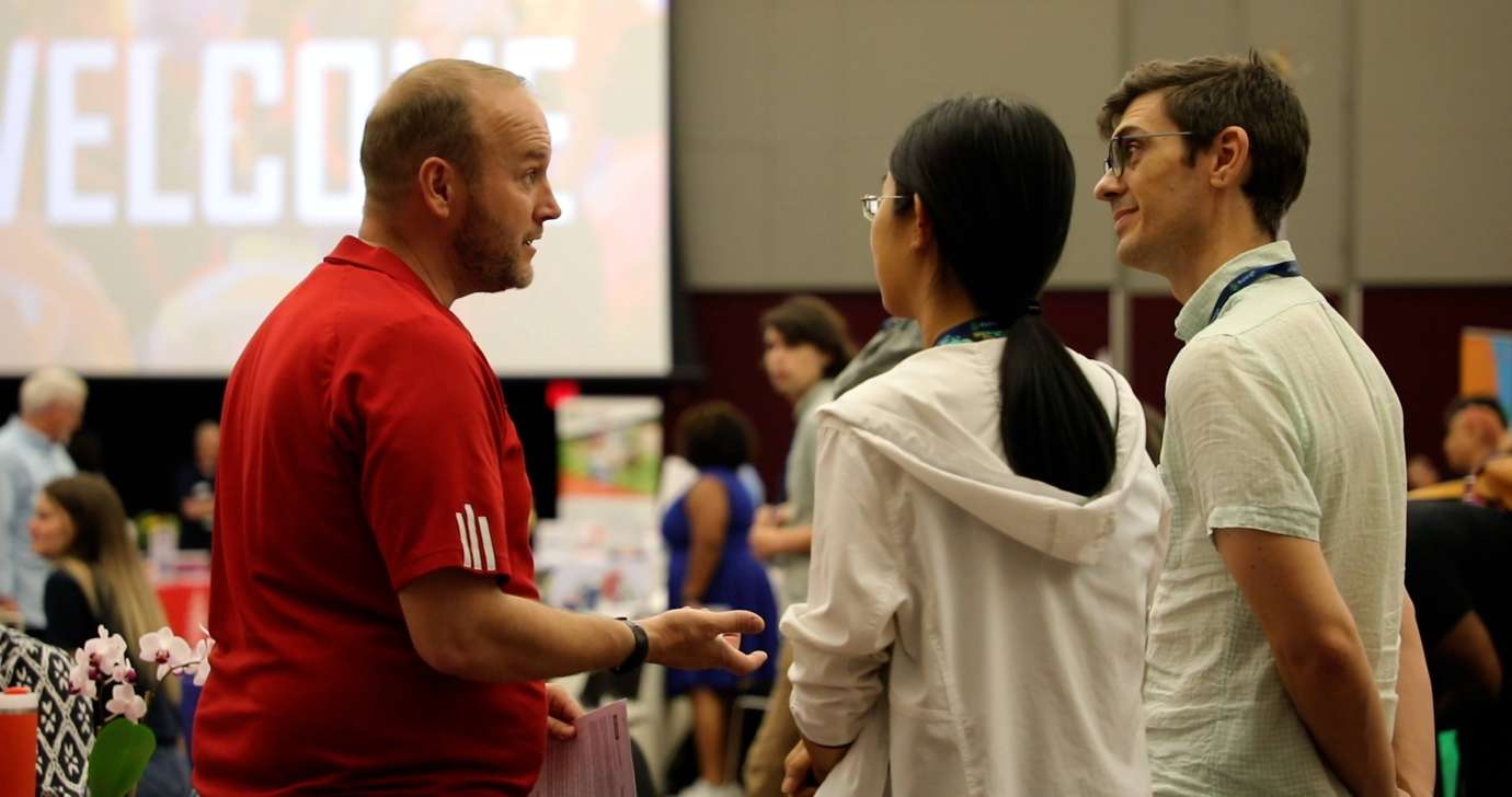 Man and woman talk to gentleman in red shirt hosting a table in the expo hall