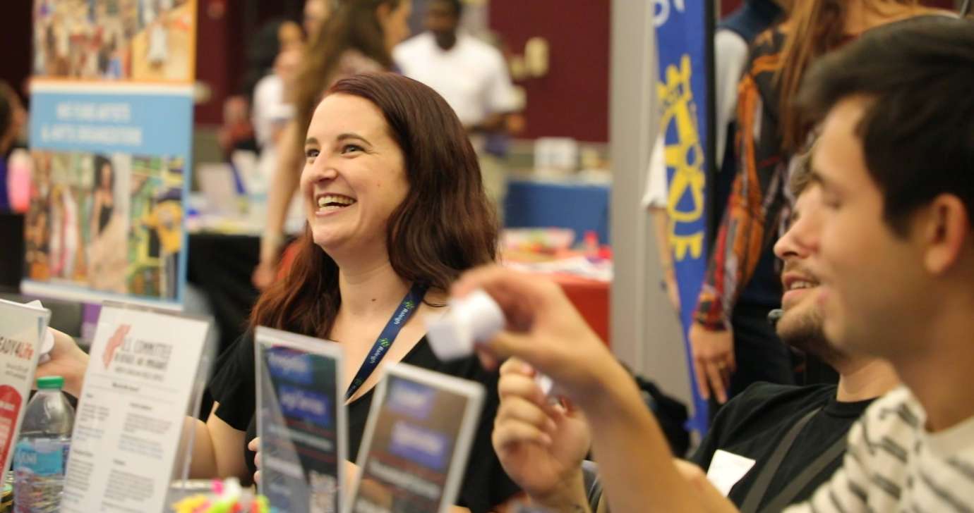 Young woman with brown hair smiling behind her table while talking to resident out of frame