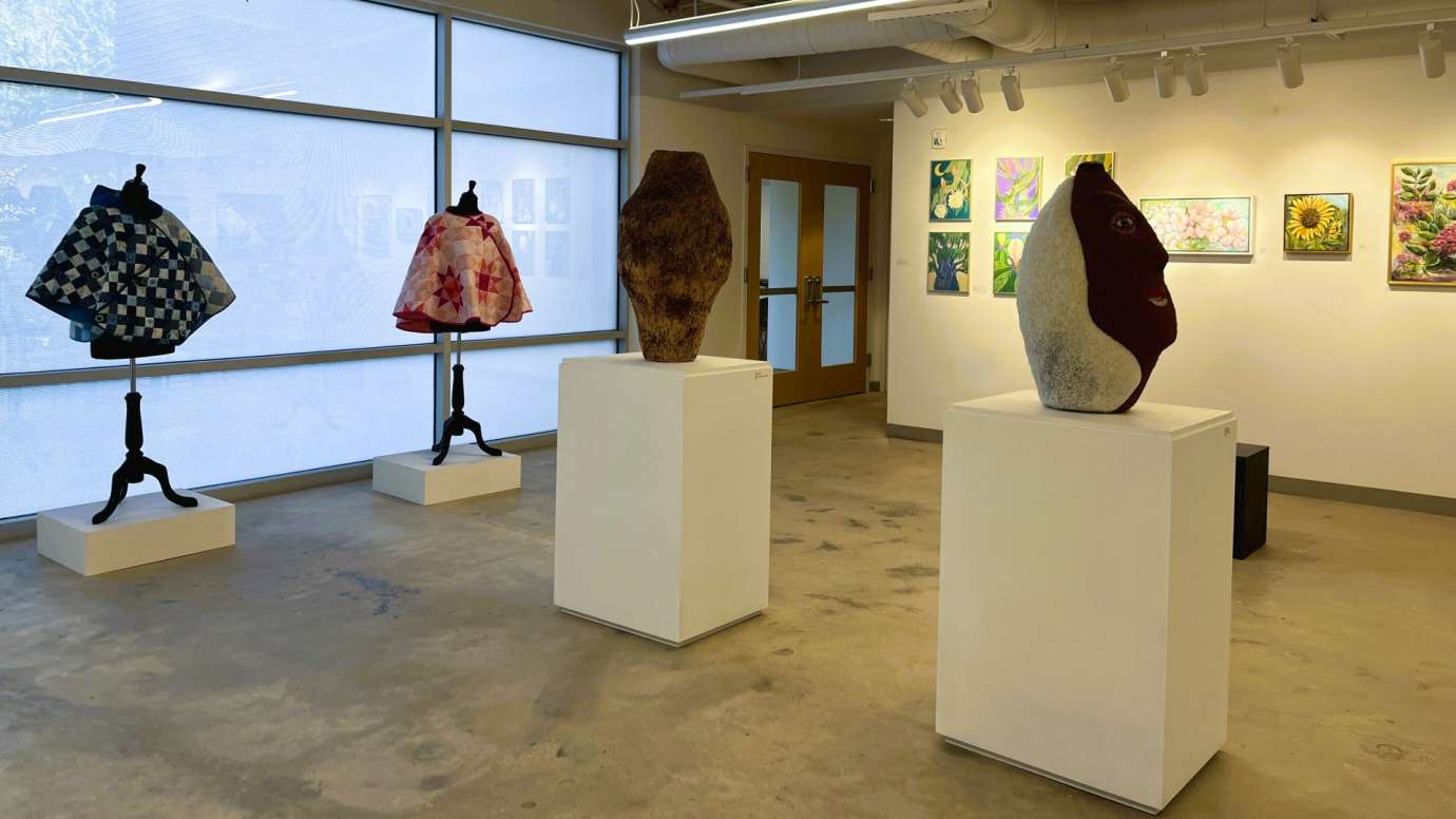 Gallery with figurative sculpture and dress forms with quilted capes in front of artwork on a wall. 