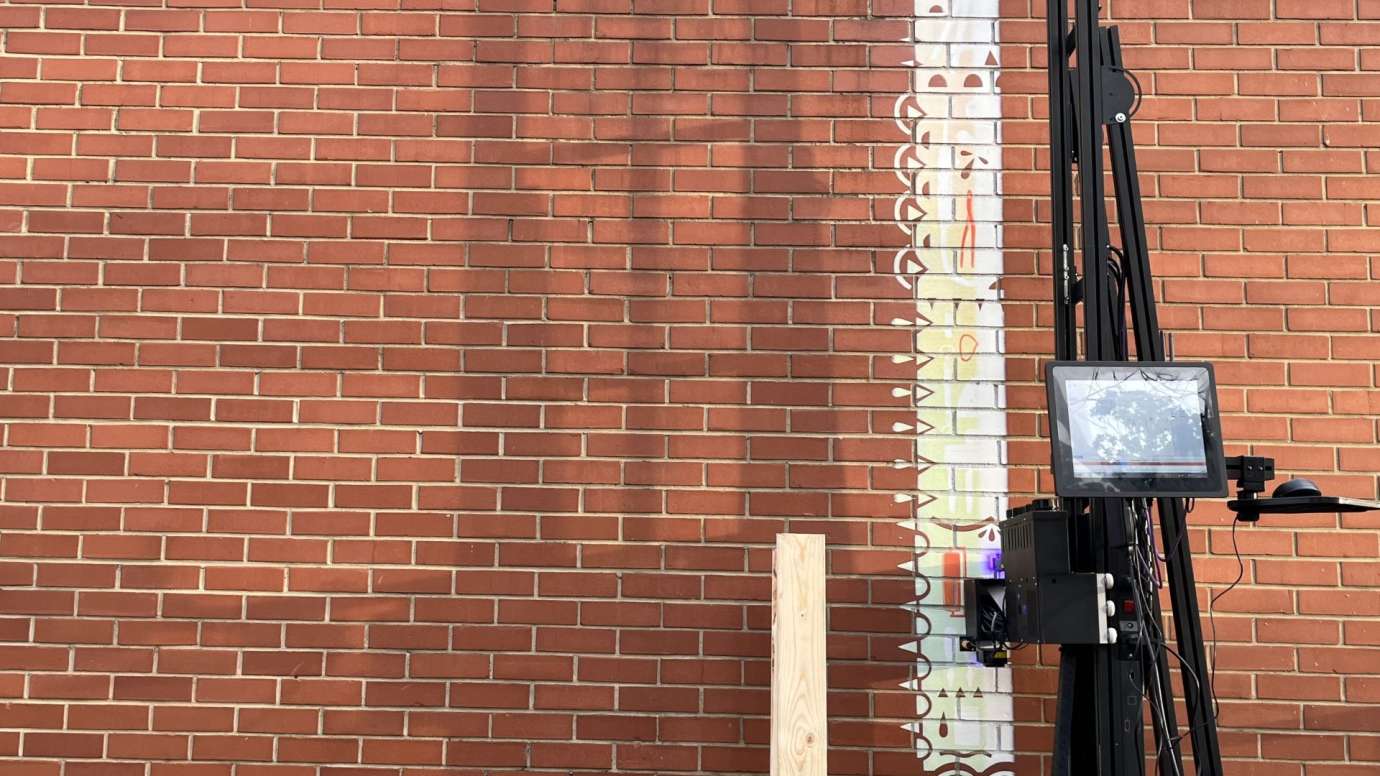 Tall black printer applying white paint to a brick wall to create a mural.