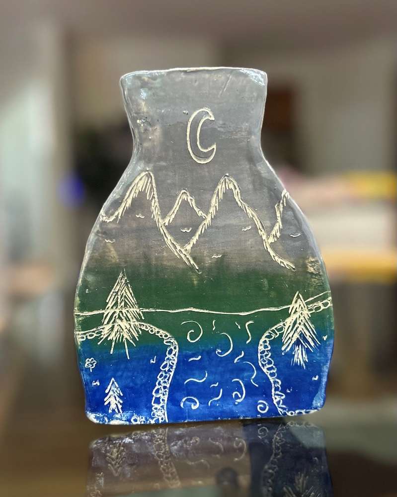 a piece of pottery with blue and green colors and etchings showing moutains, the moon, water and trees