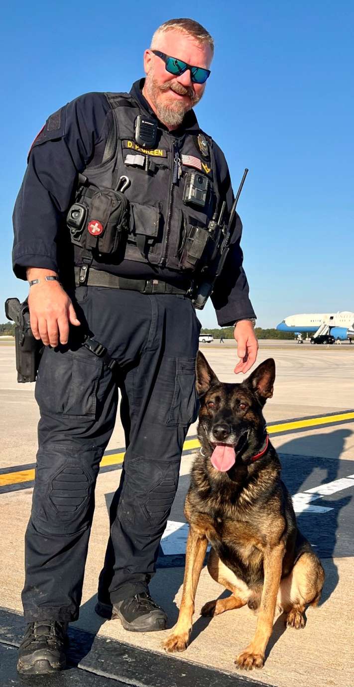 A sunny day, Officer Green stands with Hugo by his side. Hugo's tongue is sticking out. 