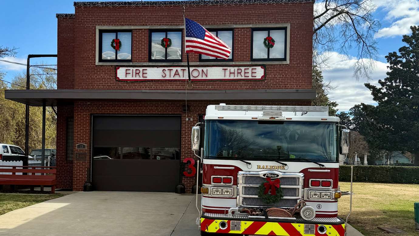 The brick building with "Fire Station Three" sign above the two garage bays is decorated for Christmas with window wreaths. A fire engine sits outside the garage bay with a wreath on front grill. 