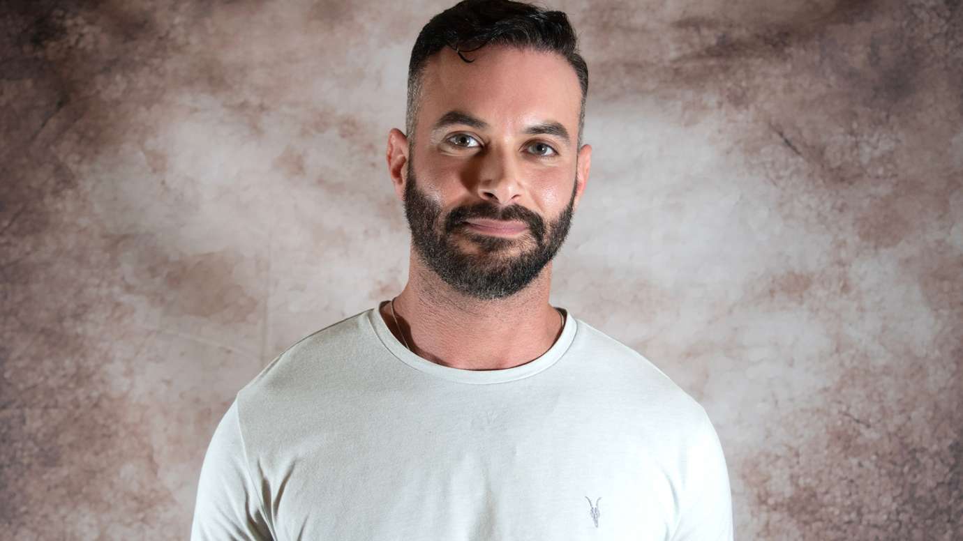 Portrait of man with dark hair, mustache and beard in a cream colored shirt, and slightly smiling at the camera