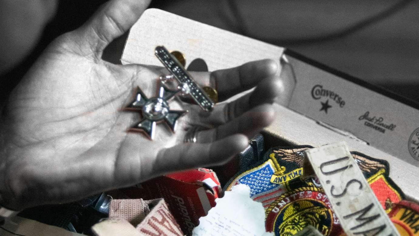 black and white photo of hand holding a Marine Rifle Sharpshooter badge, with marine and various memorabilia in color in the background