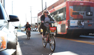 Two women riding Citrix bicycles with a GoRaleigh bus in the background