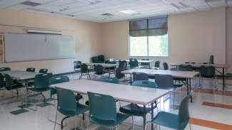 A medium sized classroom with multiple tables and chairs and a whiteboard 