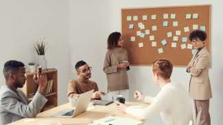Two women standing using post-it notes and wall board while collaborating with three men who are seated