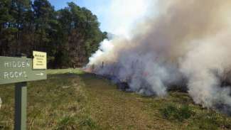 Prescribed burn at nature preserve with burning ground and smoke rising with sign on left