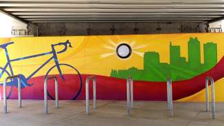 A close up photo of a mural by artist JP Jermaine Powell on wall behind metal bike racks at Raleigh Union Station. The mural features a bicycle, a sun painted around a light fixture, and a silhouette of the city skyline with colorful wavy lines.