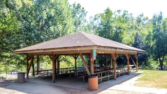 Wooden shelter surrounded by green trees with picnic tables underneath